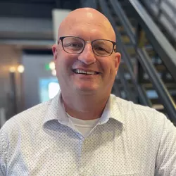 Photo of a bald man with glasses wearing a white button down shirt standing in front of black stairs. Community and Economic Development Director Chad Bunger